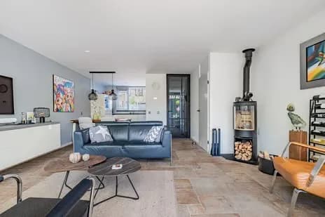 Een gewone woonkamer met een blauwe bank, een houtkachel en een open doorkijk naar de keuken